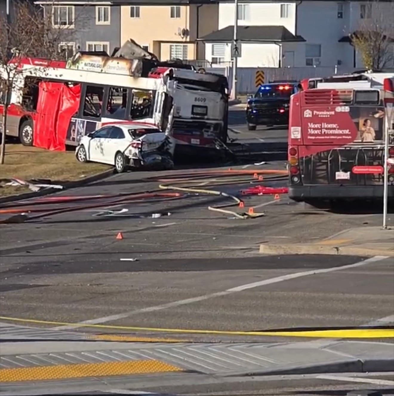 Transit buses and a damaged white car at a collision scene, with emergency vehicles visible in the background.