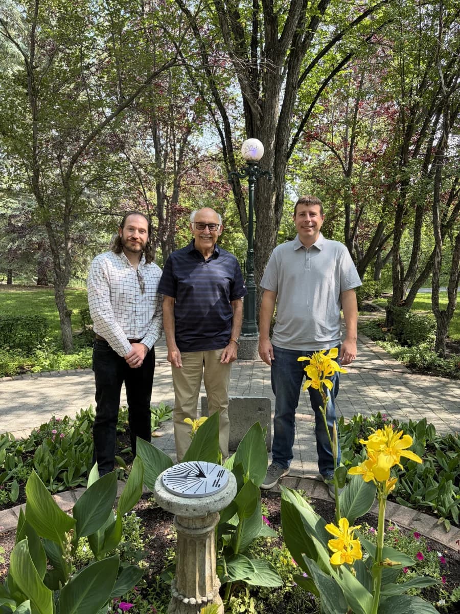 Three people standing beside a garden sundial surrounded by flowers and mature trees.